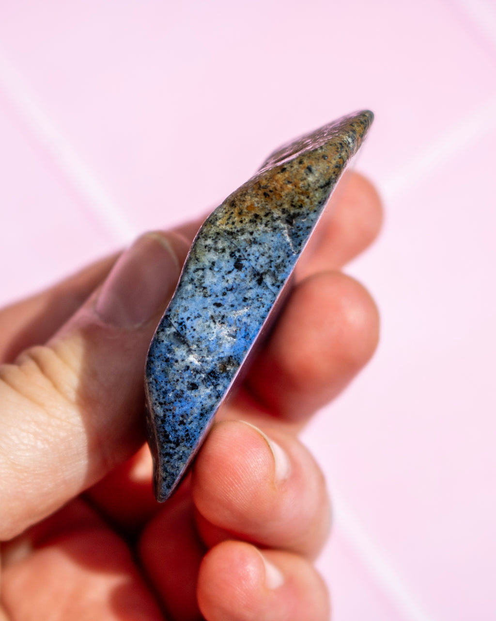 A hand holding a polished blue crystal pendant against a soft pink background.