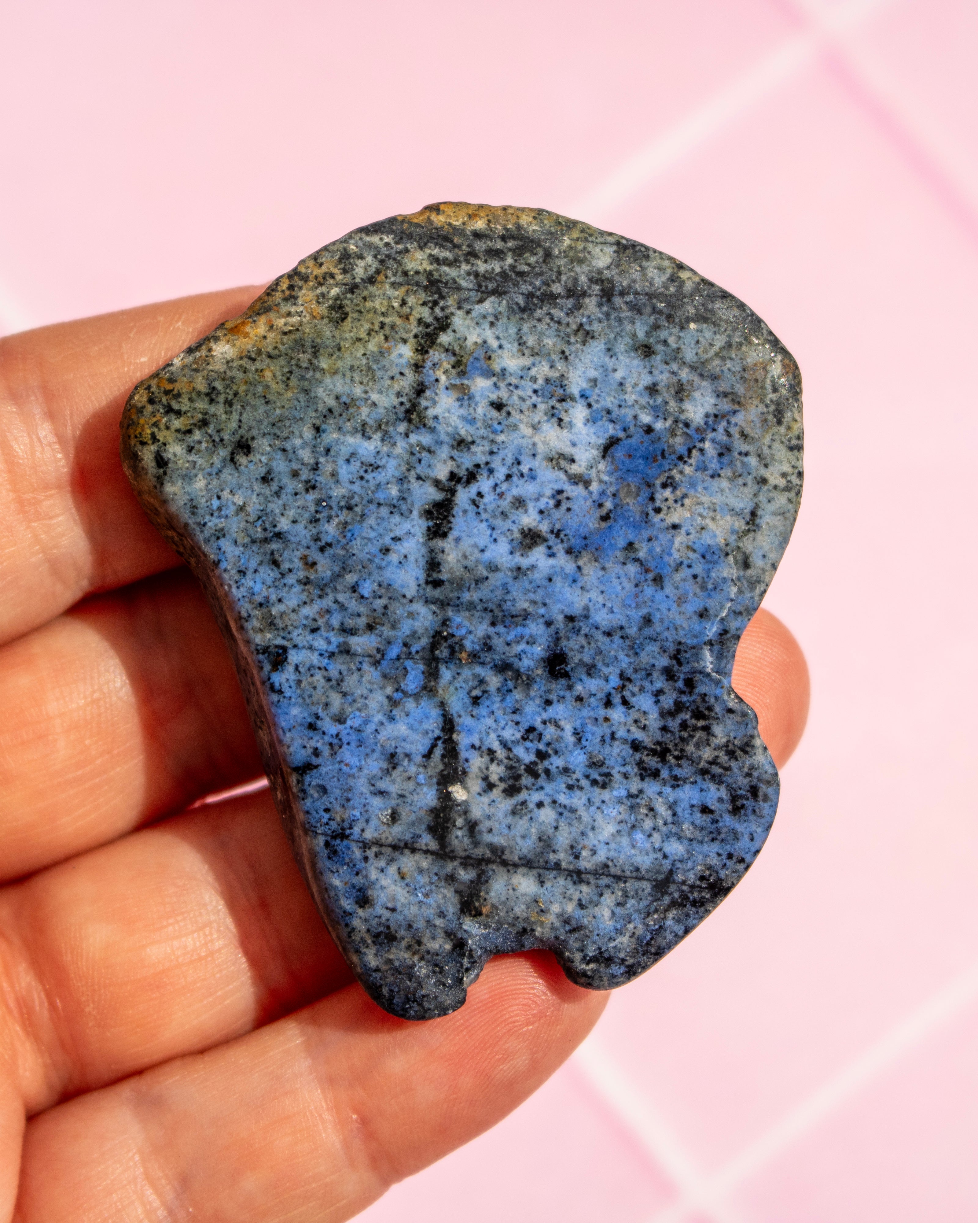 A hand holding a polished blue stone with black and brown speckling, set against a soft pink background.