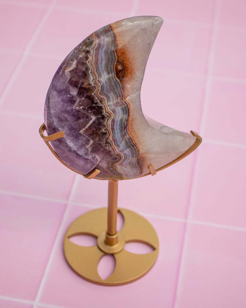 A crescent moon-shaped crystal cluster, likely amethyst, displayed on a gold-colored metal stand against a pink tiled background.