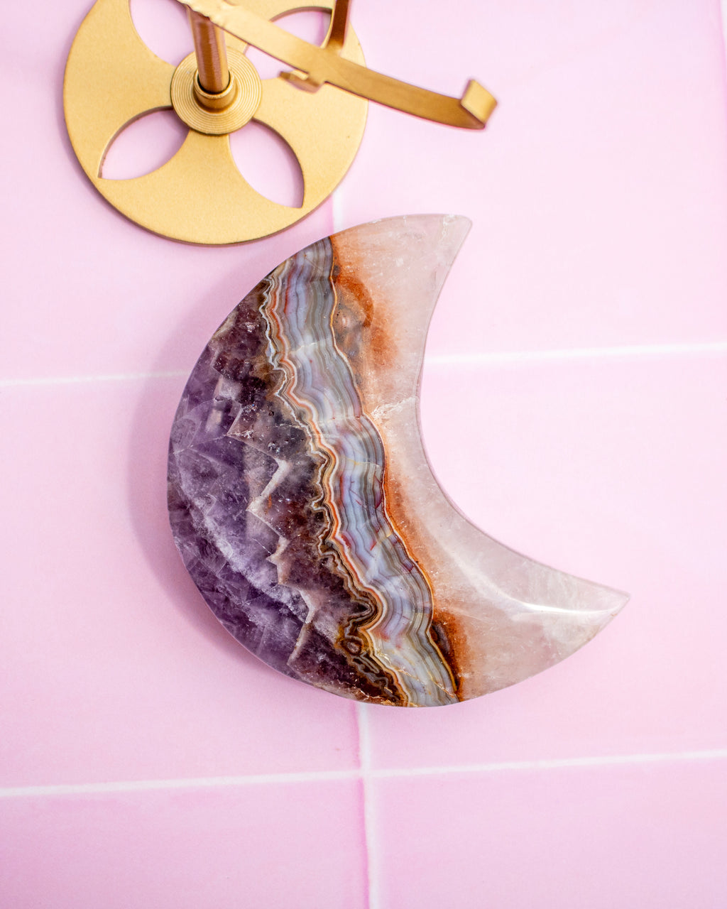 A crescent moon-shaped stone with a purple, brown, and white agate geode pattern, shown on a pink tiled wall next to a gold wall hook.