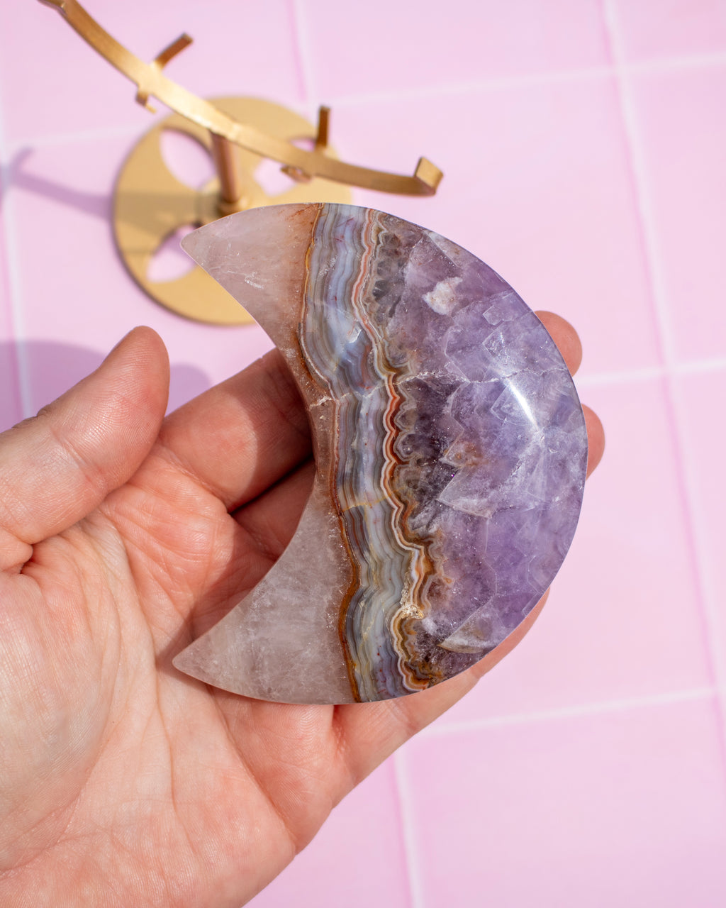 A hand holds a polished crescent moon-shaped crystal stone with purple, lavender, gray, and brown agate-like banding, set against a pink tiled background with a gold-colored object partially visible in the upper left.