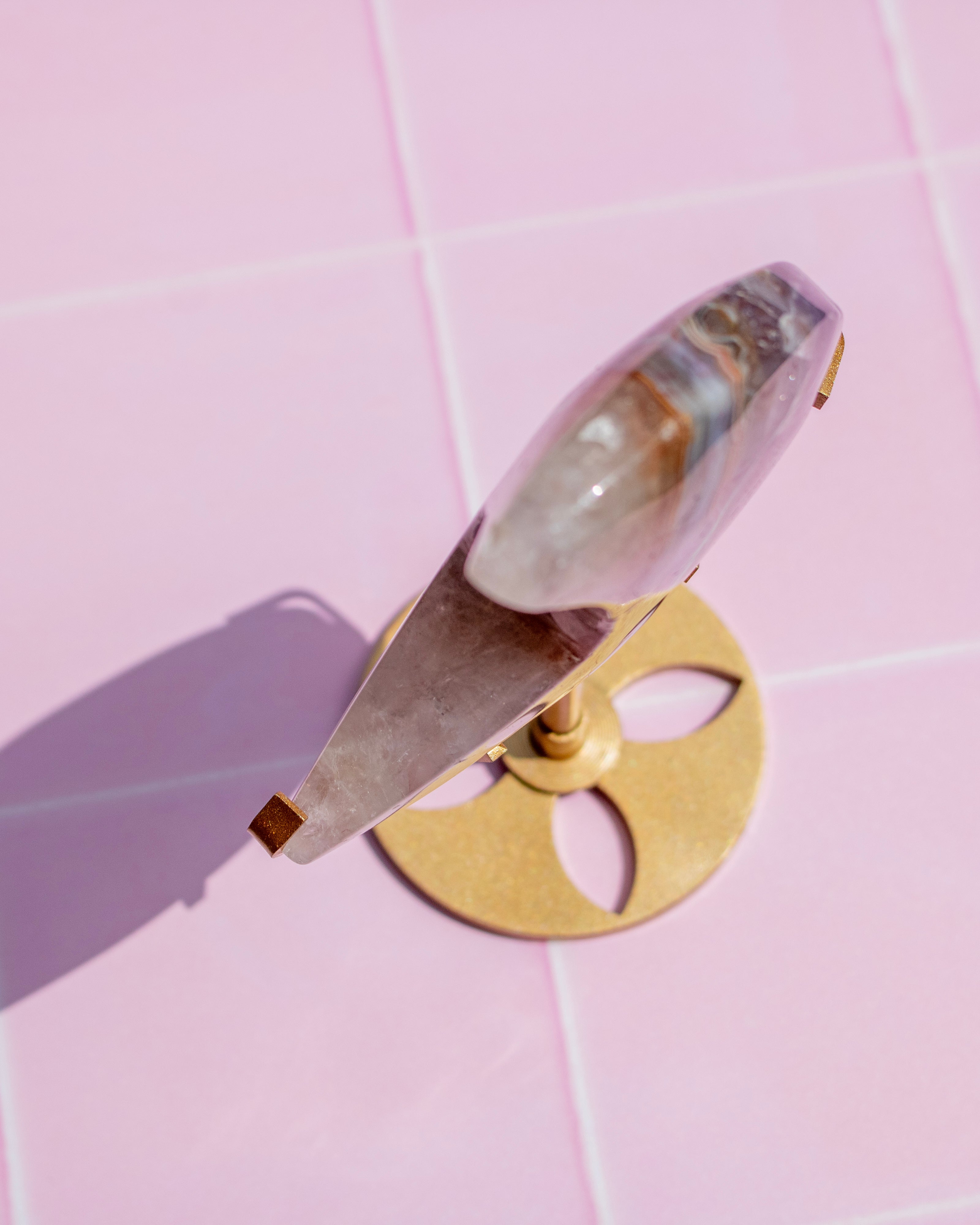 A clear quartz crystal on a gold metal stand, photographed from an overhead angle on a light pink tiled background.