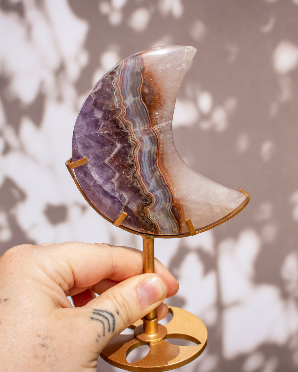A hand holds a crescent moon-shaped crystal cluster made of purple, gray, and brown banded stone on a gold-colored metal stand against a soft gray background with leaf shadows.