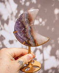 A hand holds a crescent moon-shaped crystal cluster made of purple, gray, and brown banded stone on a gold-colored metal stand against a soft gray background with leaf shadows.