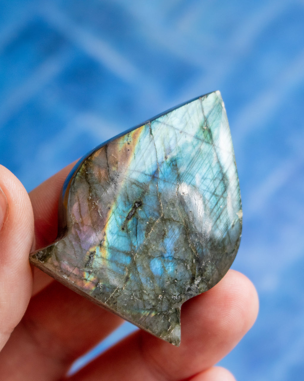 A hand holding a polished labradorite stone pendant that displays a shimmering blue, green, and gold iridescent play-of-color, set against a soft, out-of-focus blue background.