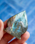 A hand holding a polished labradorite stone pendant that displays a shimmering blue, green, and gold iridescent play-of-color, set against a soft, out-of-focus blue background.