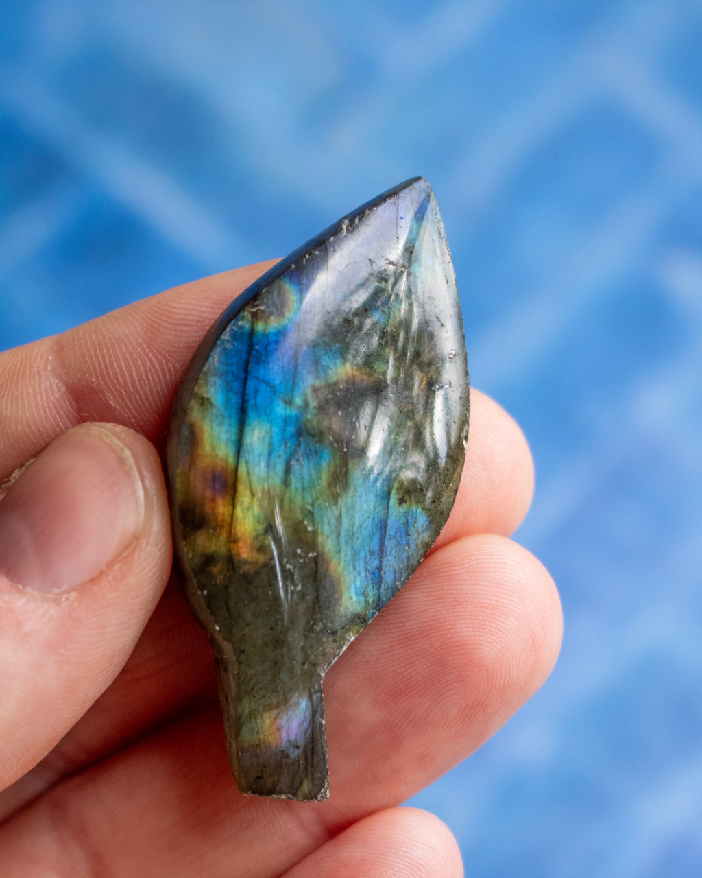 A hand holding a polished labradorite stone pendant, showcasing its characteristic blue, green, and gold iridescent flashes against a blurred blue background.