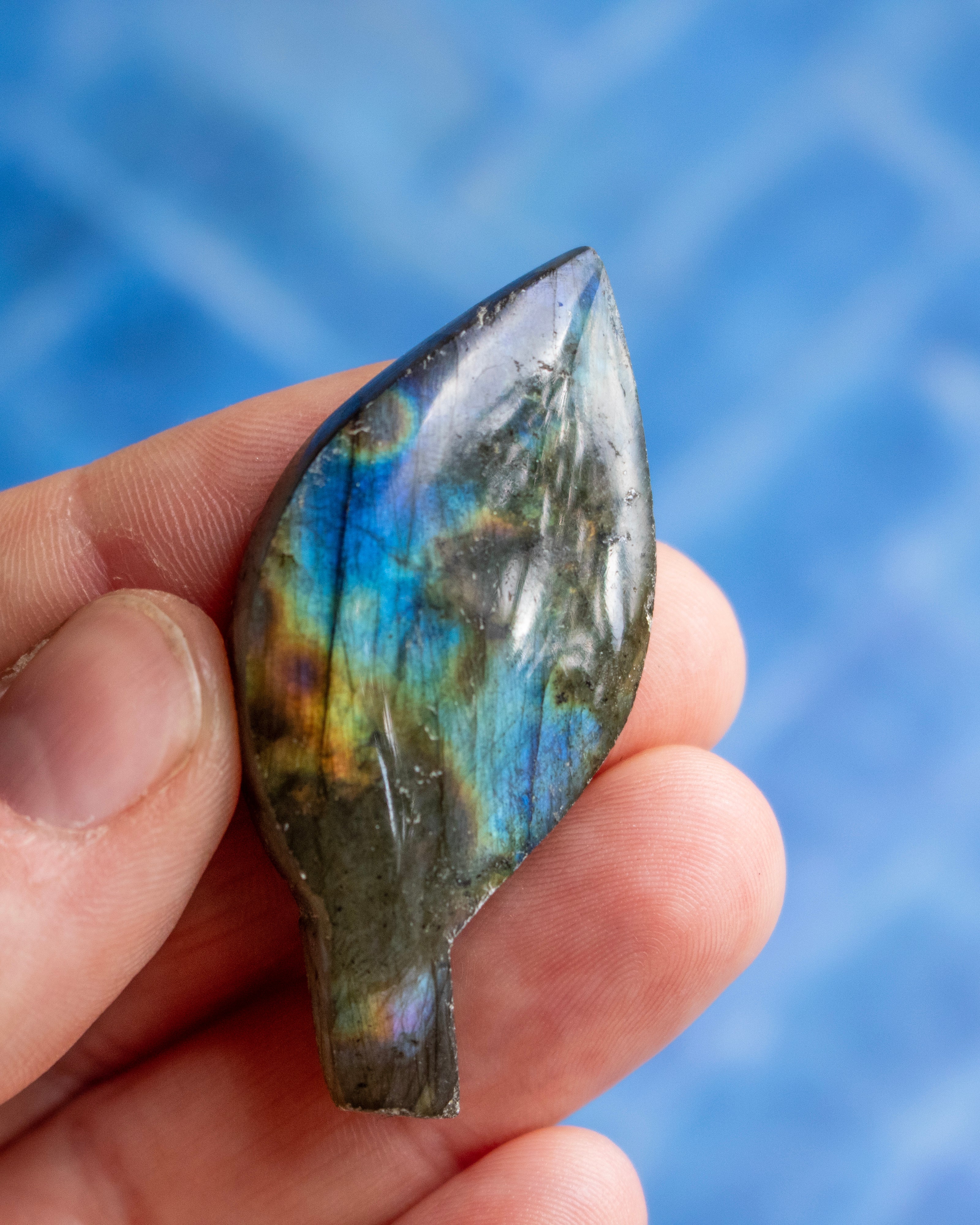 A hand holding a polished labradorite stone pendant, showcasing its characteristic blue, green, and gold iridescent flashes against a blurred blue background.
