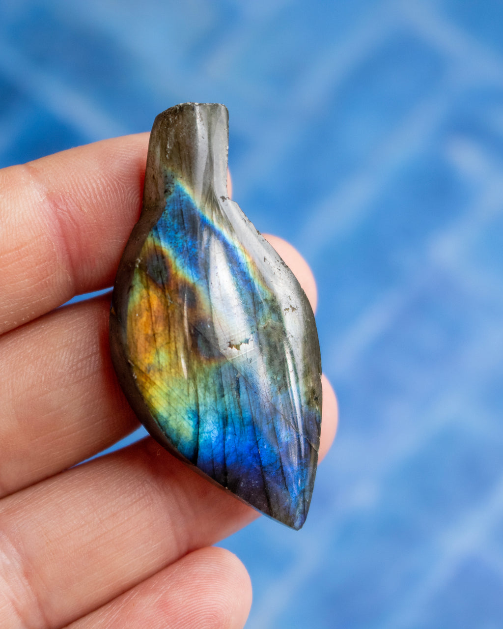 A hand holding a polished labradorite stone pendant with vibrant blue, green, and gold iridescent flashes, set against a textured blue background.