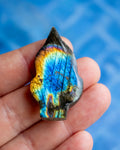 A hand holding a polished labradorite stone teardrop-shaped crystal, showcasing its vibrant blue, yellow, and orange iridescent flashes against a blurred blue background.