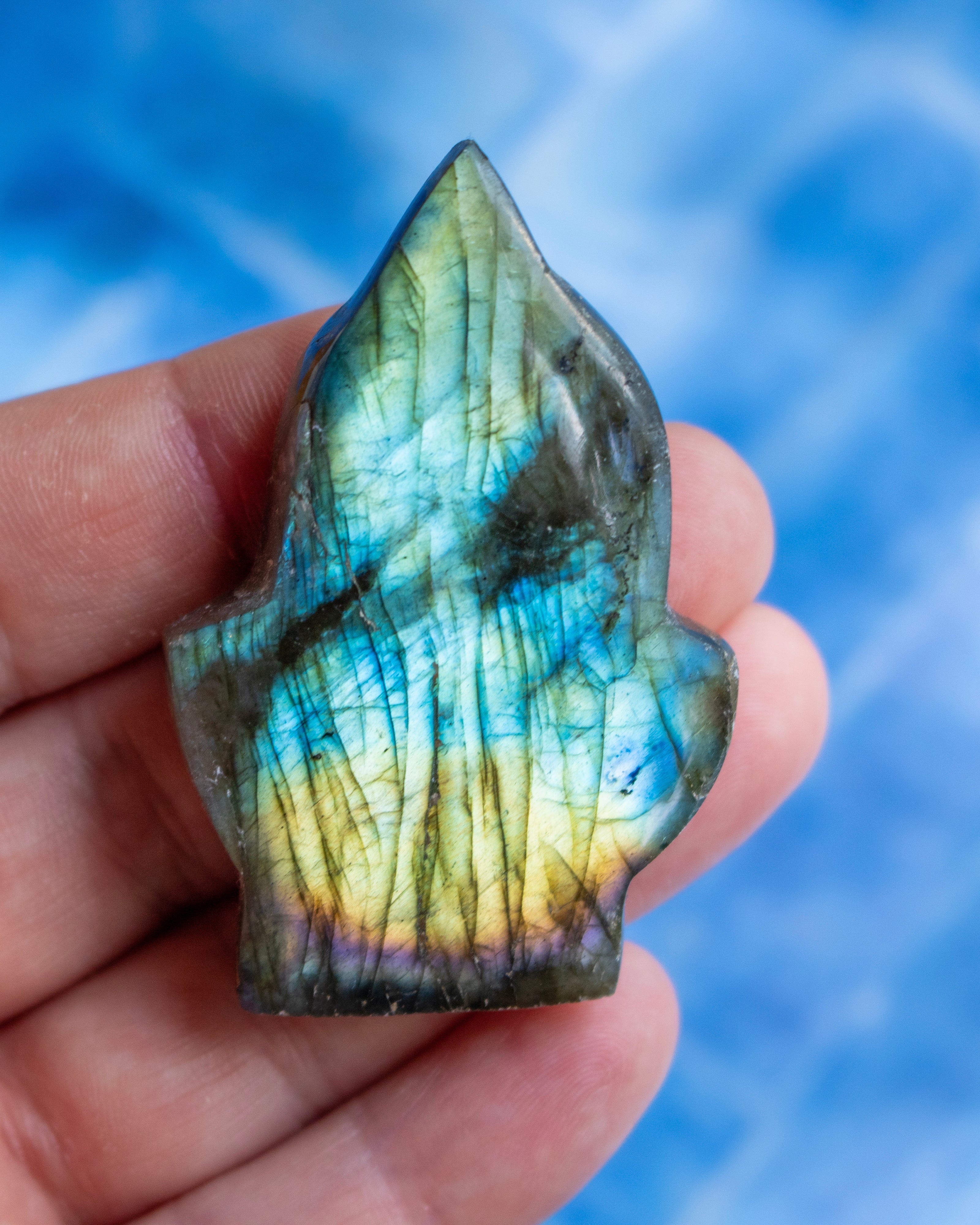 A hand holding a polished labradorite crystal point showing its characteristic blue, green, and yellow iridescent flashes, set against a blurred blue background.