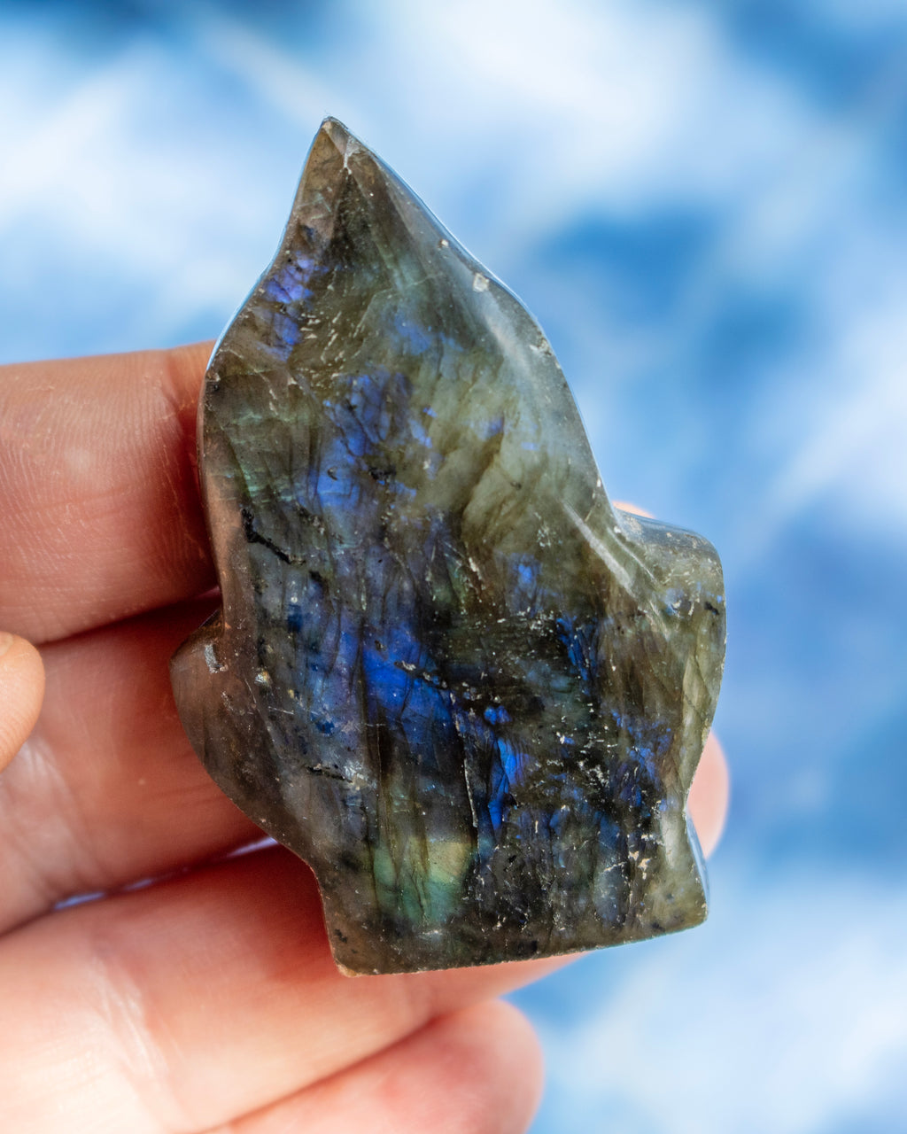 A hand holding a raw, polished labradorite crystal showing its characteristic blue and green iridescent flashes against a soft, out-of-focus blue sky background.