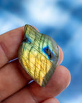 A hand holding a polished labradorite stone with a shimmering gold, green, and blue color shift against a blurred blue background.