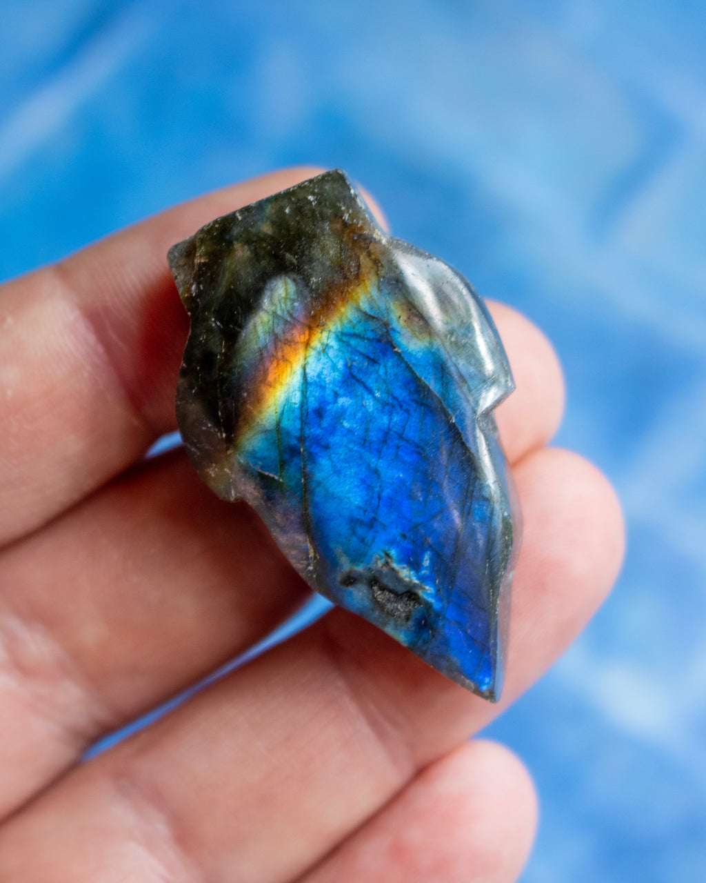 A hand holding a polished labradorite crystal showing a vibrant blue flash with an iridescent rainbow sheen, set against a soft blue background.