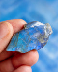 A hand holding a raw, polished blue labradorite stone against a soft, out-of-focus blue background.