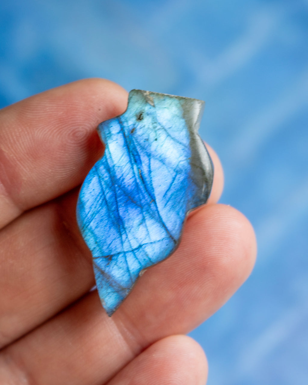 A hand holding a polished blue labradorite stone with natural gray and white flashes, set against a soft, out-of-focus blue background.