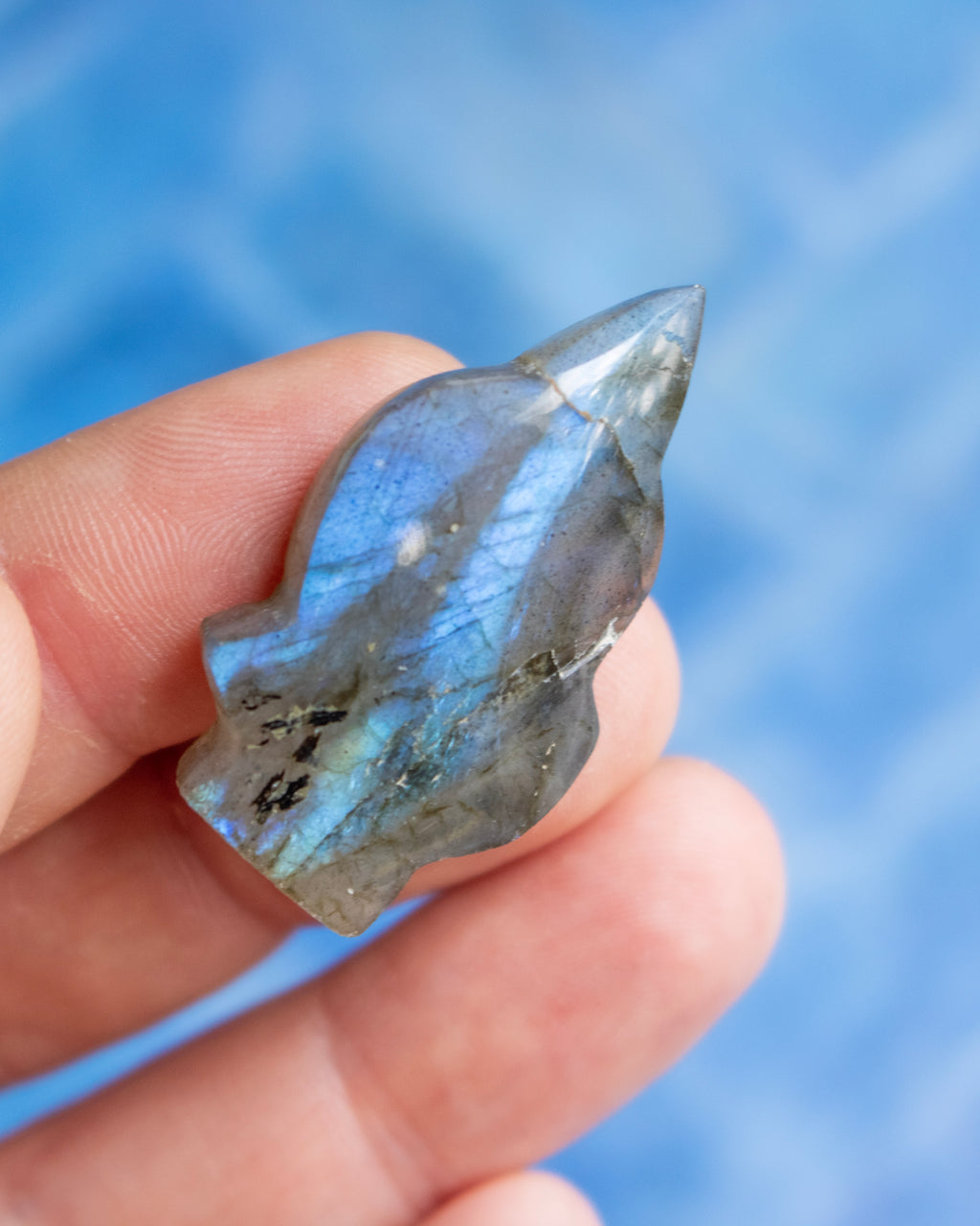 A hand holding a polished labradorite crystal point against a soft blue blurred background.