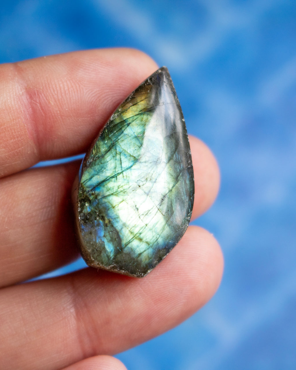 A teardrop-shaped labradorite gemstone held between fingers, showcasing its characteristic green and blue iridescent flashes against a soft blue background.