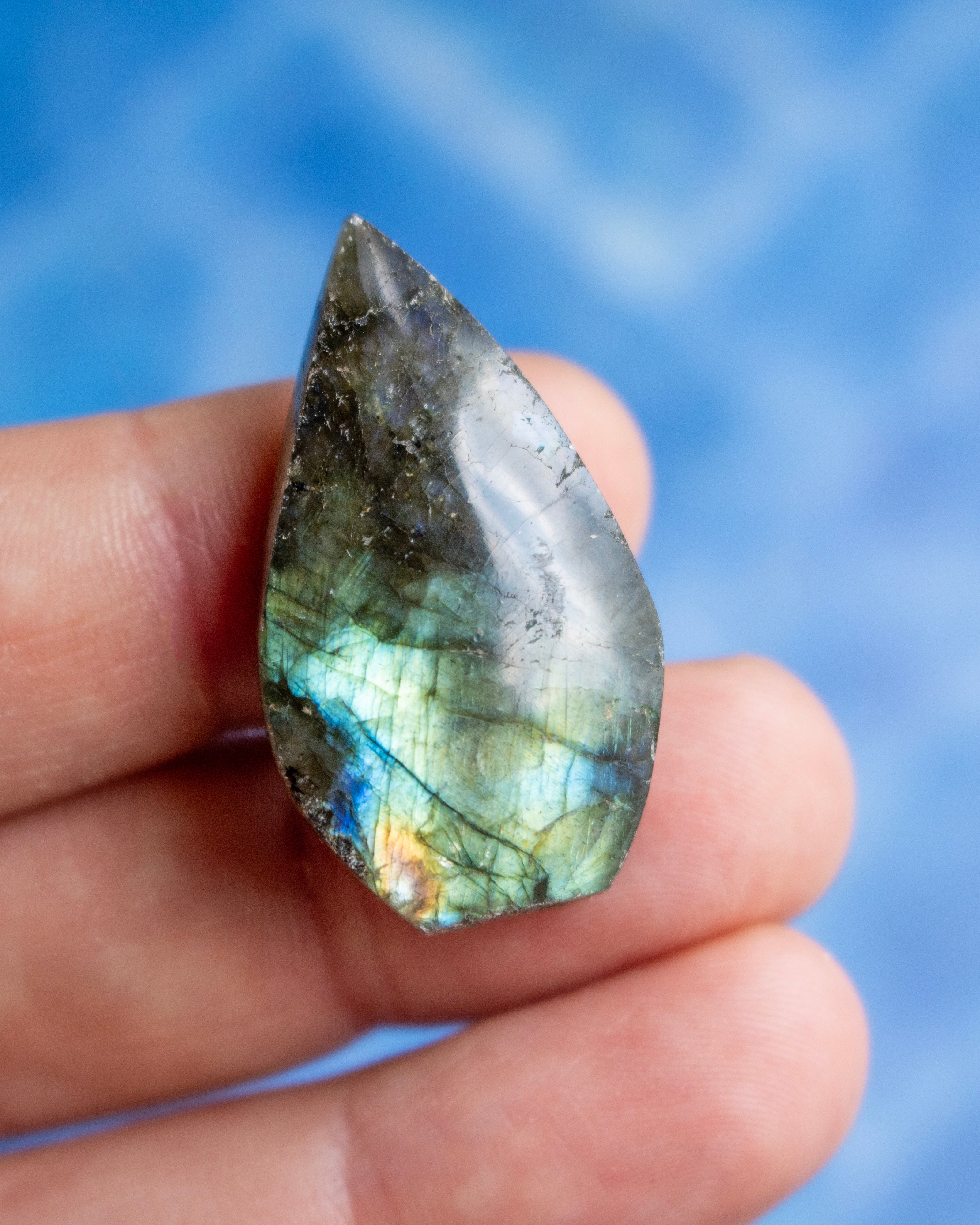 A hand holding a polished teardrop-shaped labradorite gemstone showing its characteristic green, blue, and gold iridescent flashes against a blurred blue background.