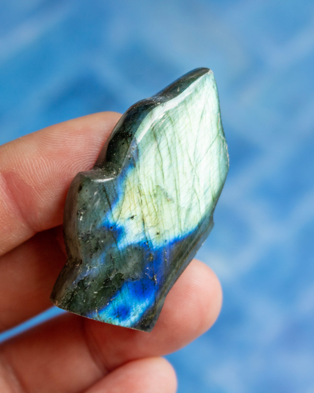 A hand holding a polished labradorite stone with a natural irregular shape, showing a color-shifting play-of-color with green, blue, and white flashes against a blurred blue background.