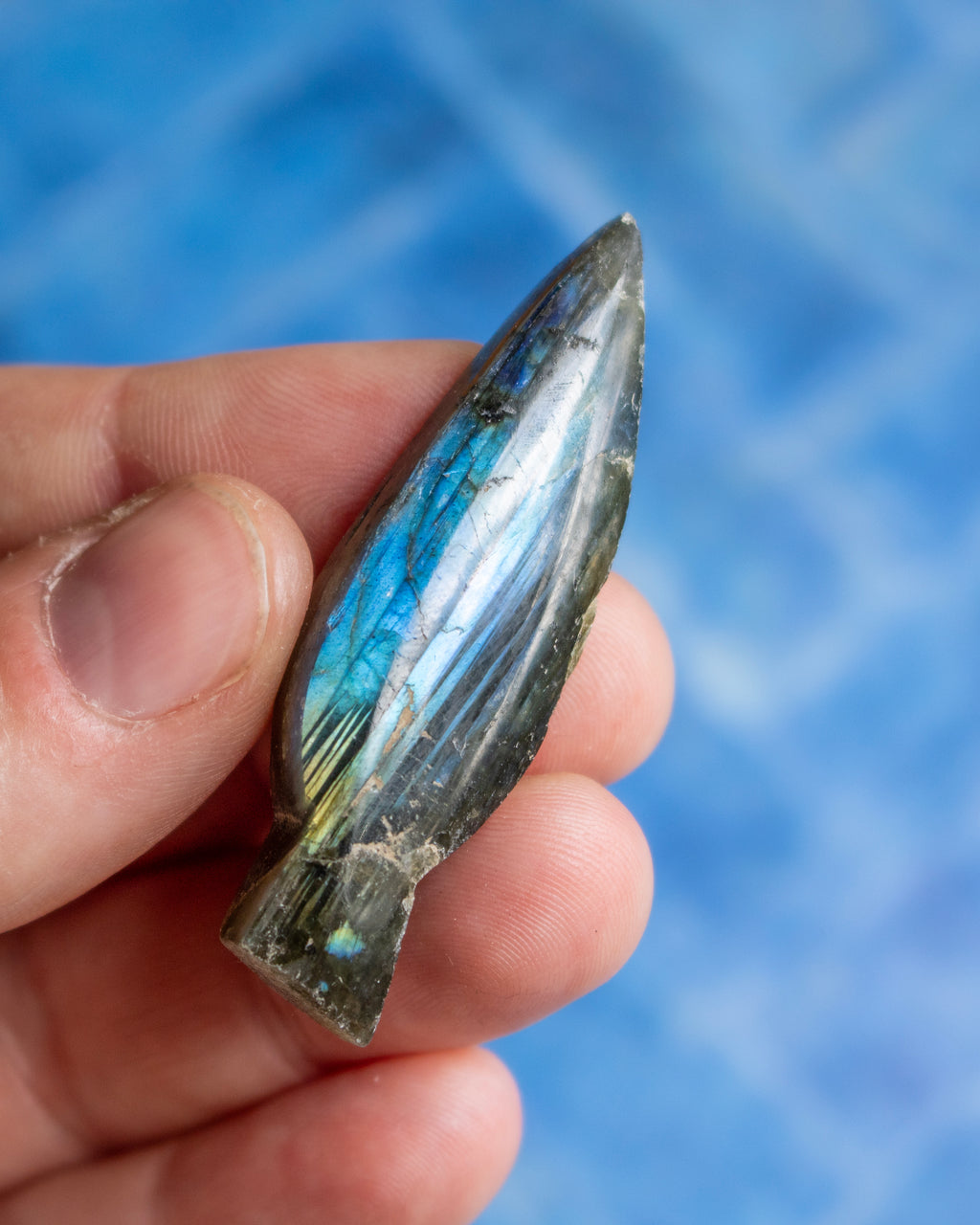 A hand holding a polished labradorite gemstone pendant with its characteristic blue and green iridescent flashes, set against a blurred blue background.