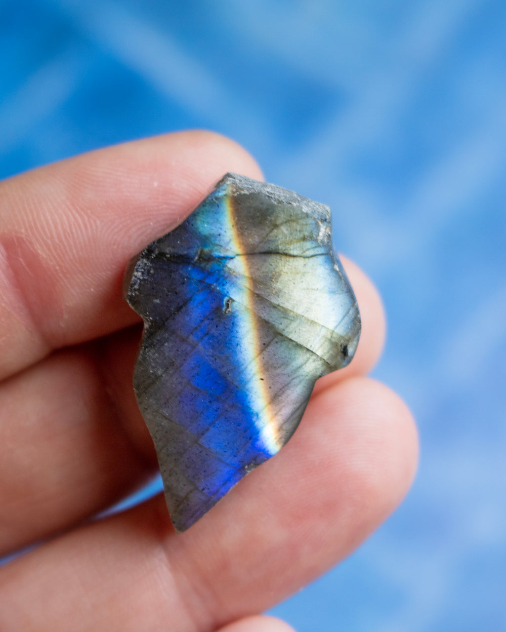 A hand holding a raw, rough piece of labradorite gemstone that displays a vibrant blue and green iridescent flash, set against a soft, out-of-focus blue background.