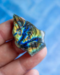 A hand holding a raw, rough-cut labradorite crystal showing vibrant blue, green, and gold iridescent flashes, set against a soft, out-of-focus blue background.