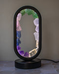 An oval-shaped table lamp with a black metal frame and base, featuring a glowing interior lined with colorful crystal points including green, pink, blue, purple, and clear quartz, set against a neutral gray wall.