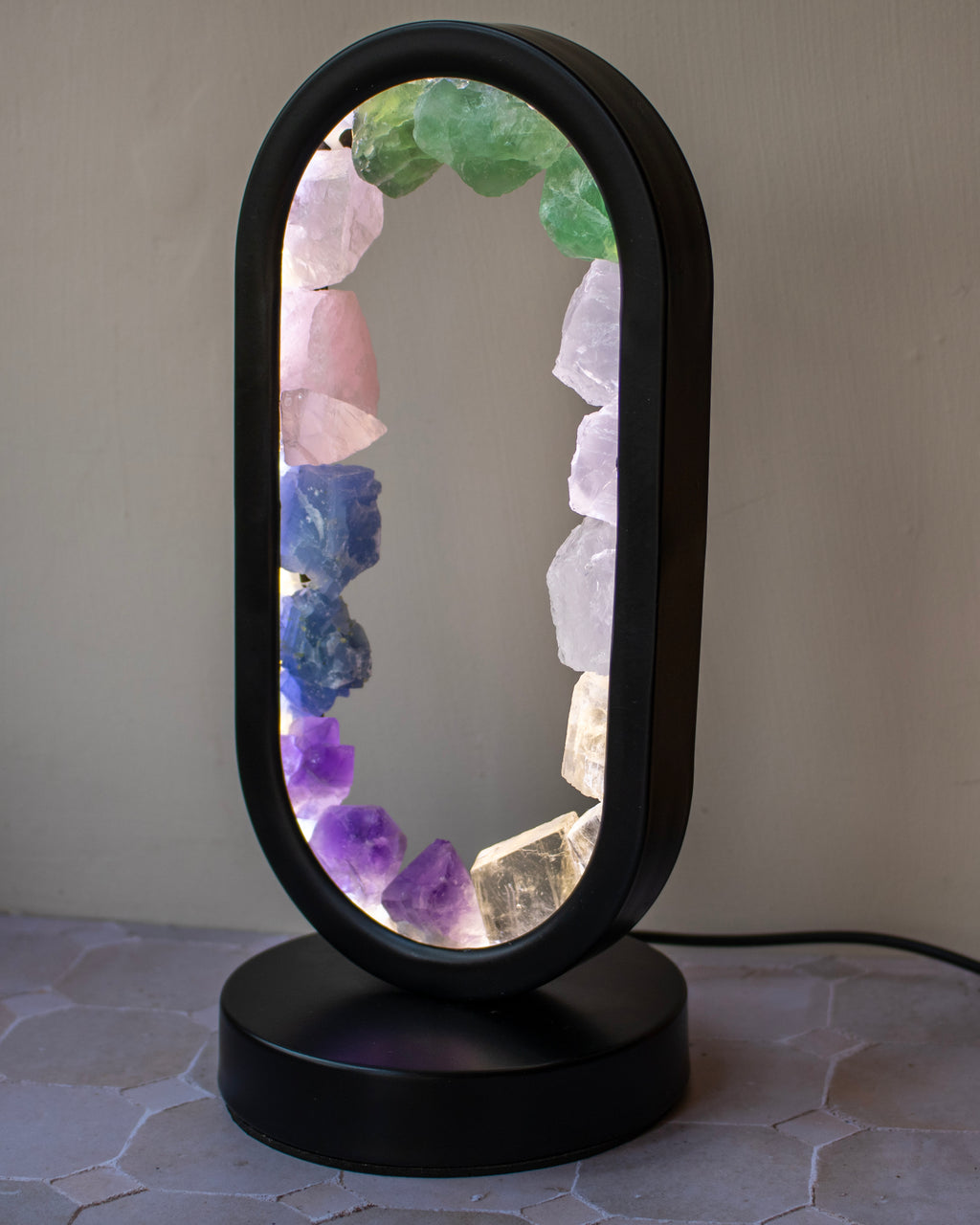 An oval-shaped table lamp with a black plastic frame and base, featuring a glowing interior lined with colorful crystal points including green, pink, blue, purple, and clear quartz, set against a neutral gray wall.