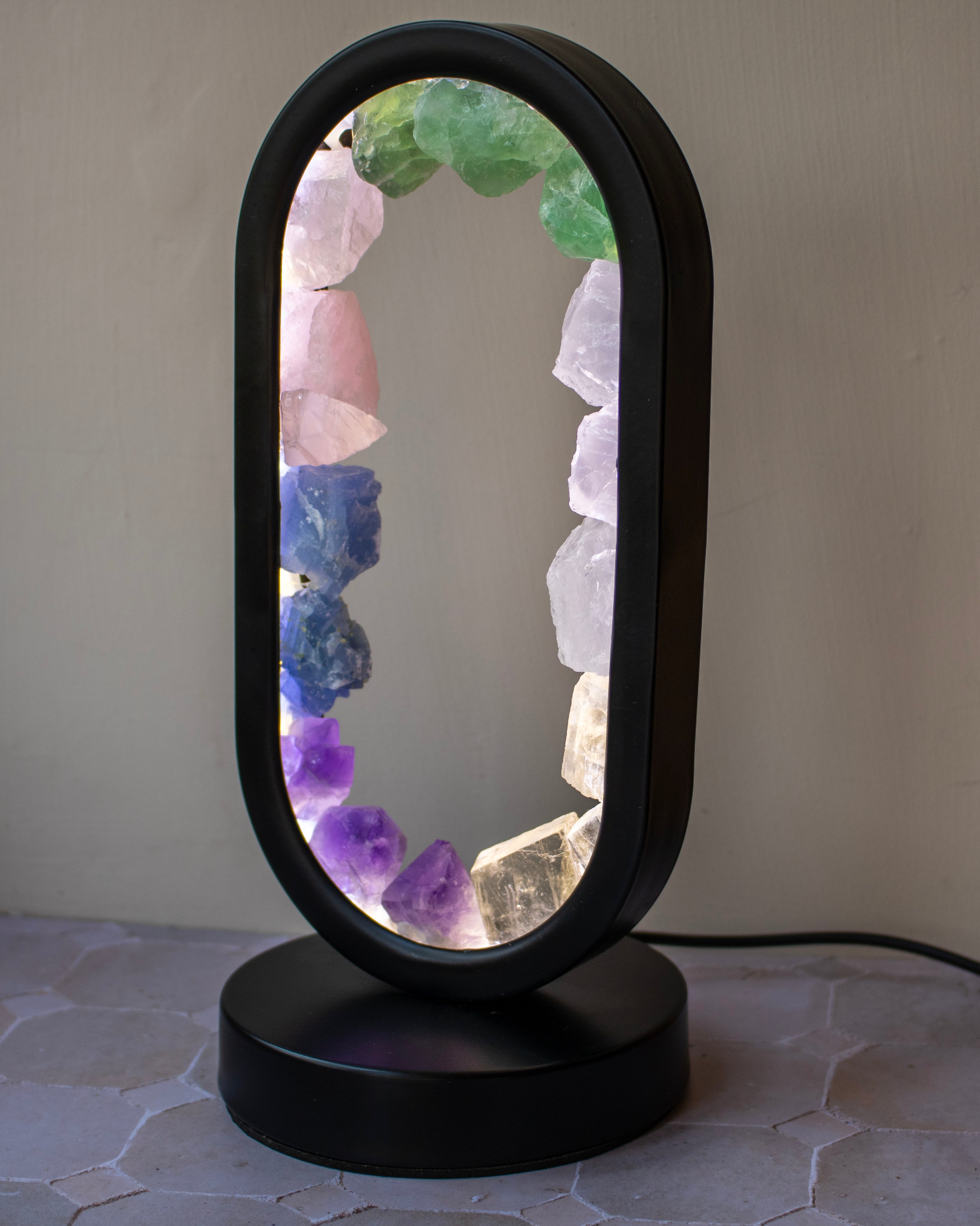 An oval-shaped table lamp with a black plastic frame and base, featuring a glowing interior lined with colorful crystal points including green, pink, blue, purple, and clear quartz, set against a neutral gray wall.