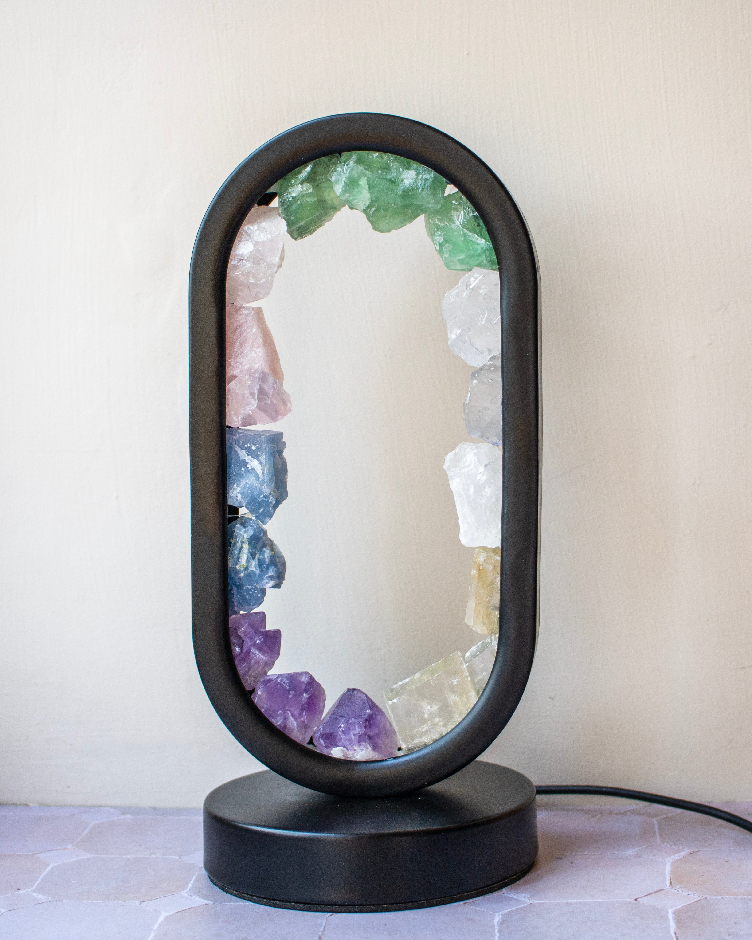A black oval table lamp with a clear center and a decorative border of raw crystal points in green, pink, blue, purple, white, and yellow, sitting on a black round base against a light beige wall.
