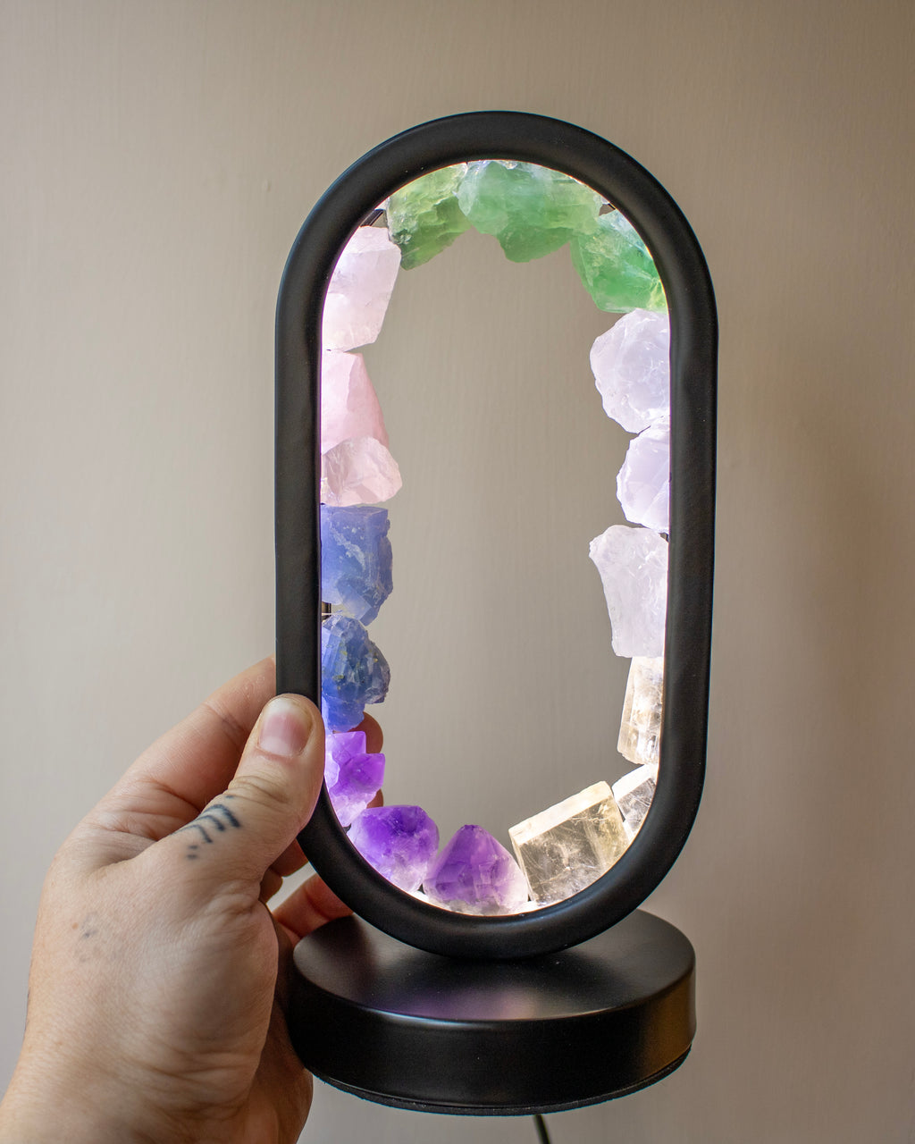 A hand holds an oval black-framed LED lamp featuring a colorful arrangement of raw crystal stones around the inner border, set against a plain beige wall.