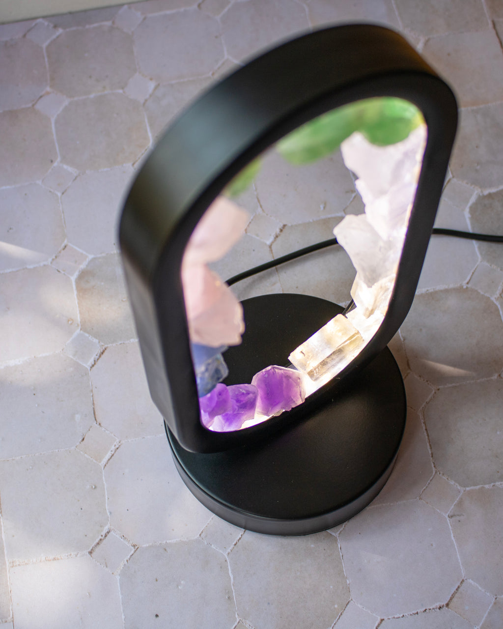 A black LED crystal lamp with a rounded rectangular frame, shown illuminated on a light gray hexagonal tile surface. The lamp contains various colored crystals, including purple, white, and green, visible through the glass panel.