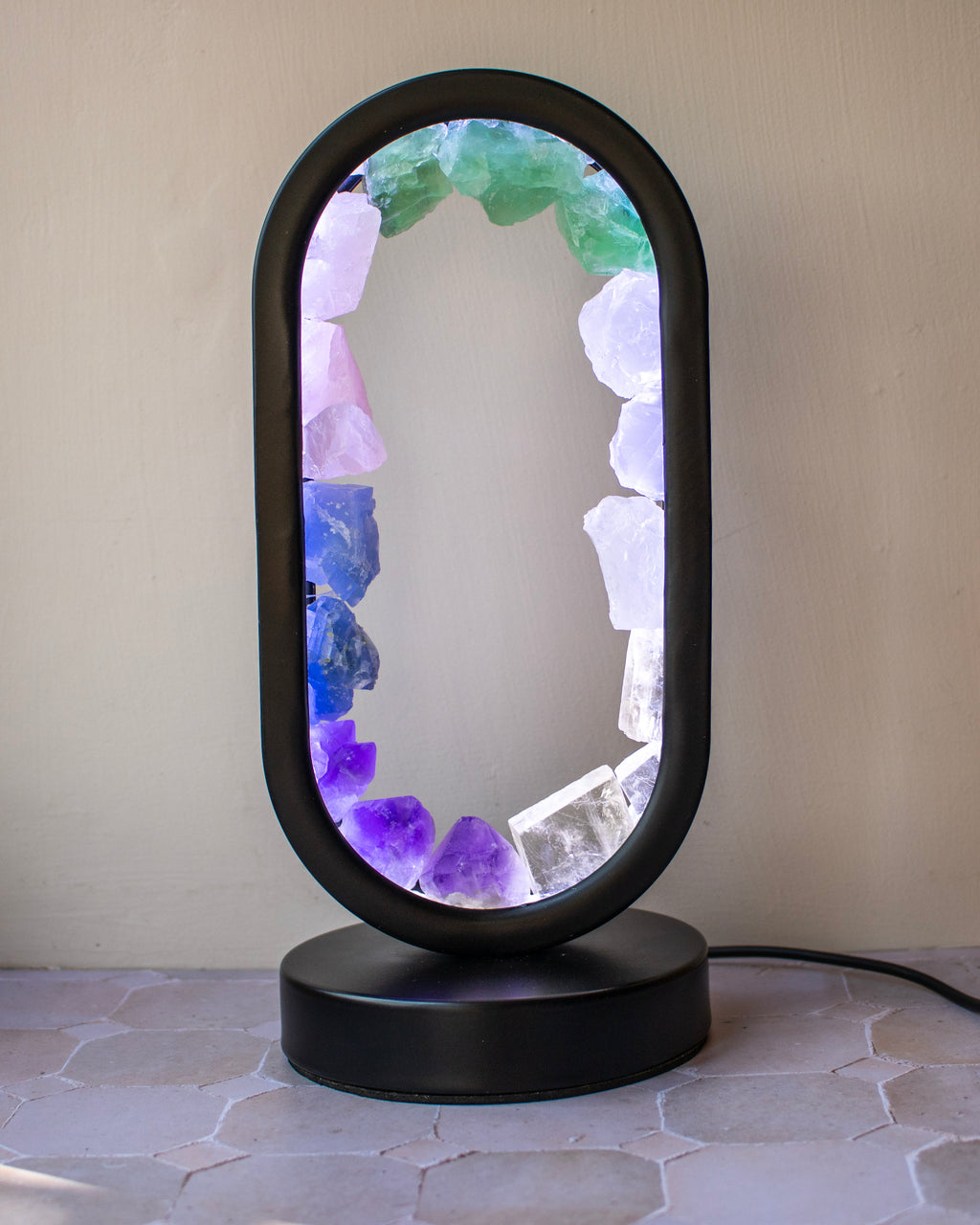 A modern oval table lamp with a matte black frame and base, featuring a glowing interior lined with raw crystals in shades of purple, blue, green, and white, set against a neutral beige wall.