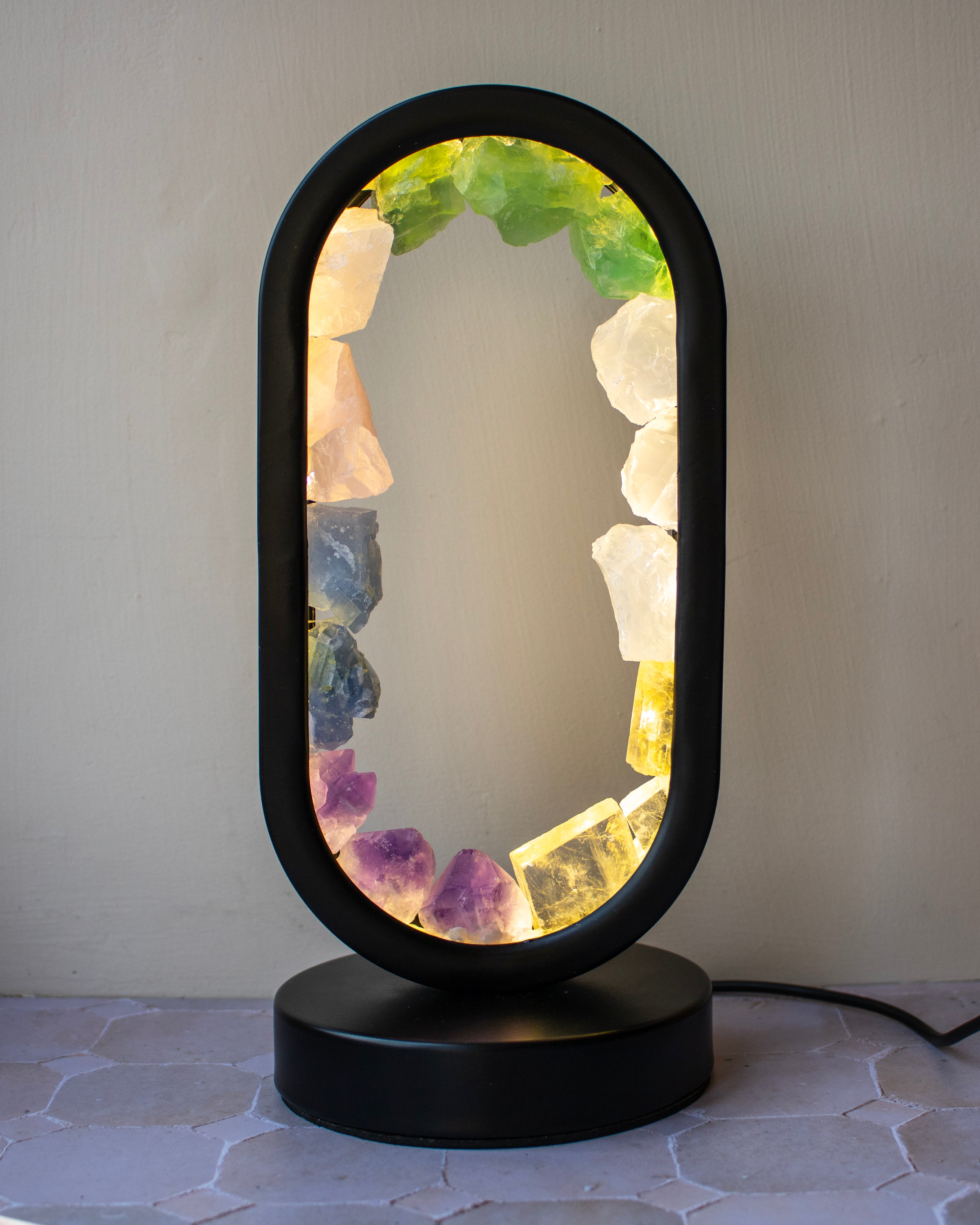 An oval-shaped black table lamp with a glowing interior lined with colorful crystal points in green, white, purple, yellow, and blue, resting on a round black base against a neutral background.