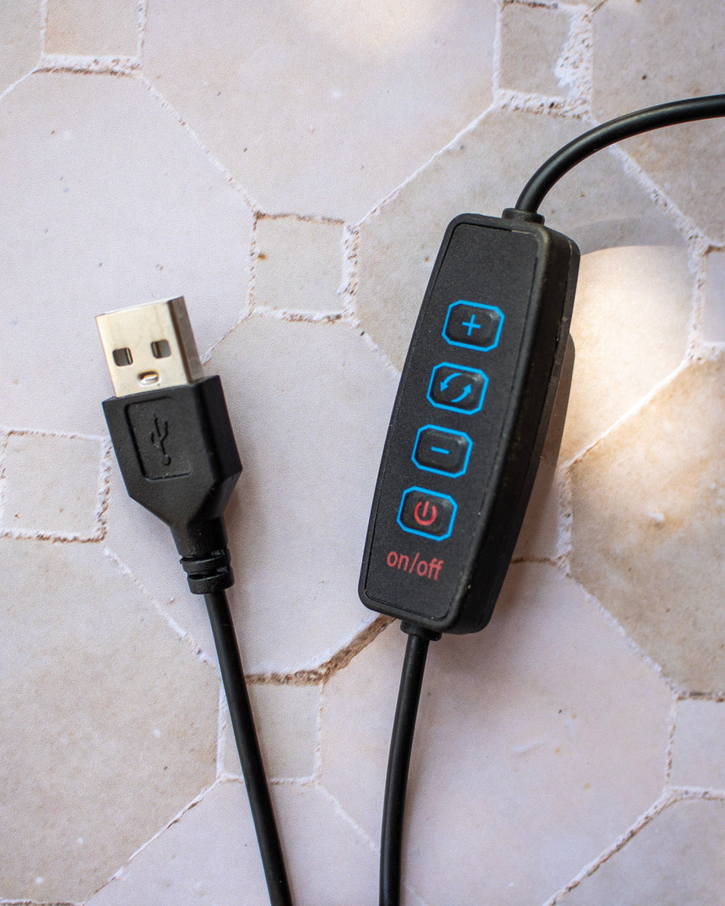 A black USB-controlled light switch with blue and red buttons, shown with its USB connector plugged in on a light beige hexagonal tile surface.