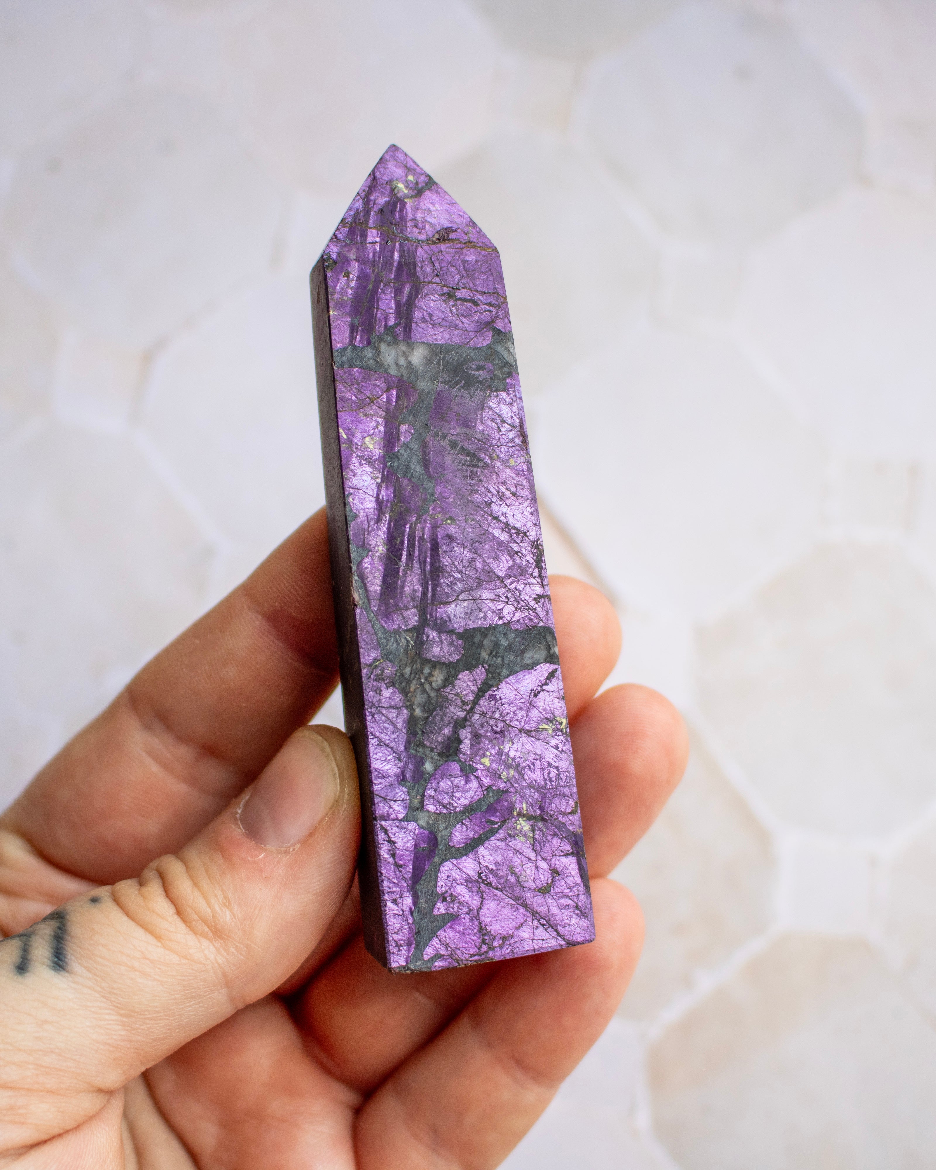A hand holding a polished purple crystal point with natural dark veining, set against a light beige stone tile background.