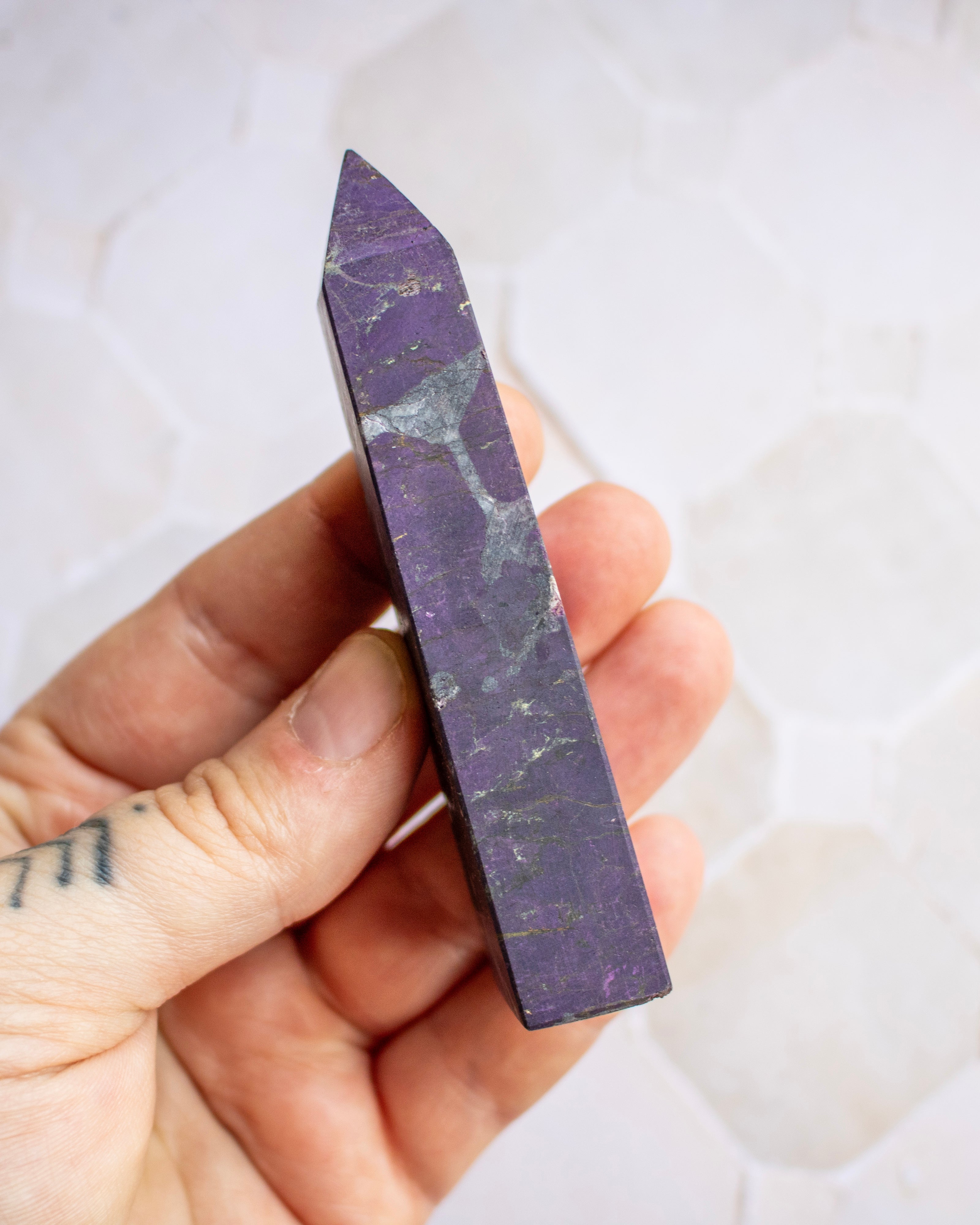 A hand holding a polished purple crystal point with natural white and gray veining, set against a light, softly blurred background.