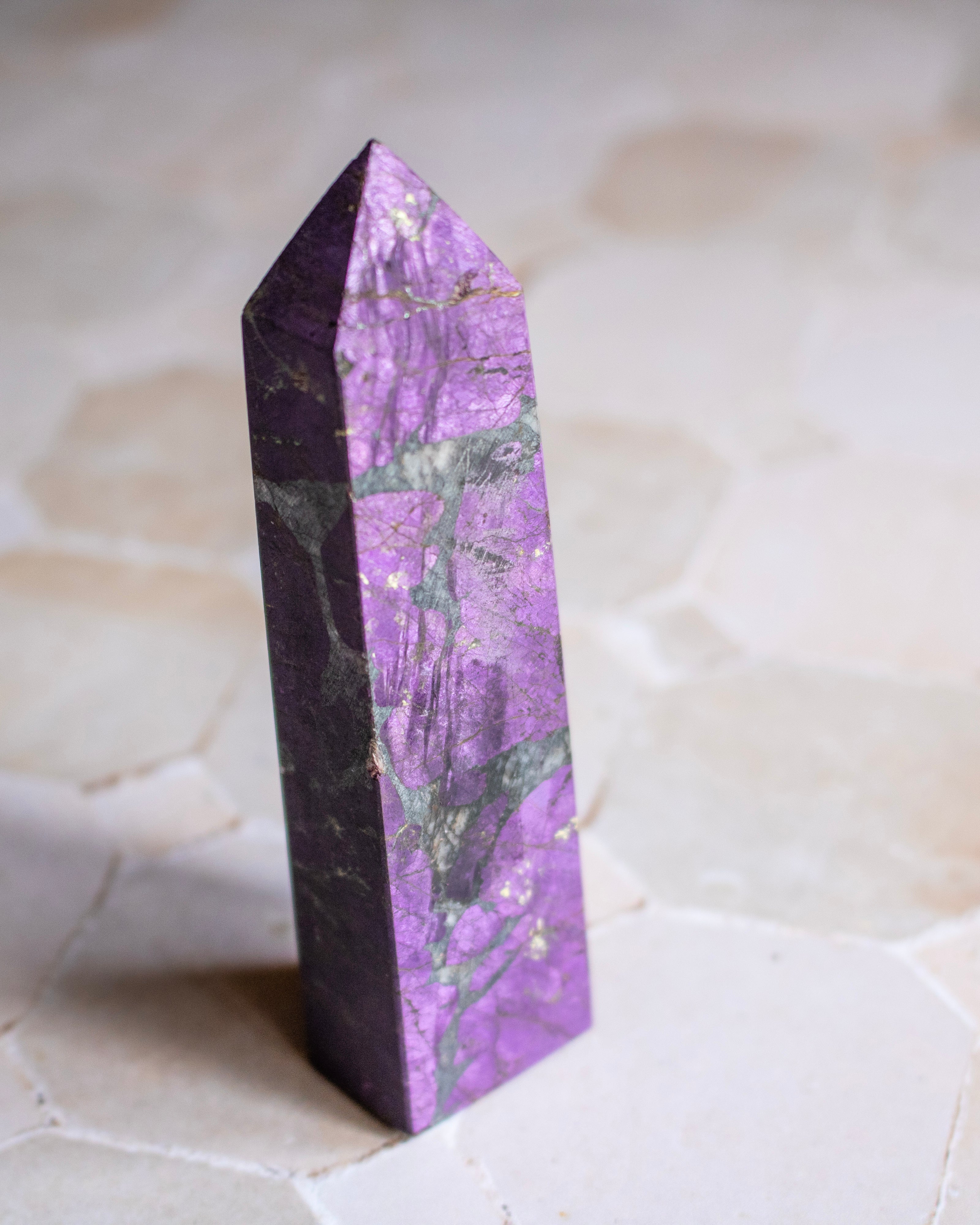A polished purple crystal tower with natural green and white veining, standing upright on a light beige stone surface.