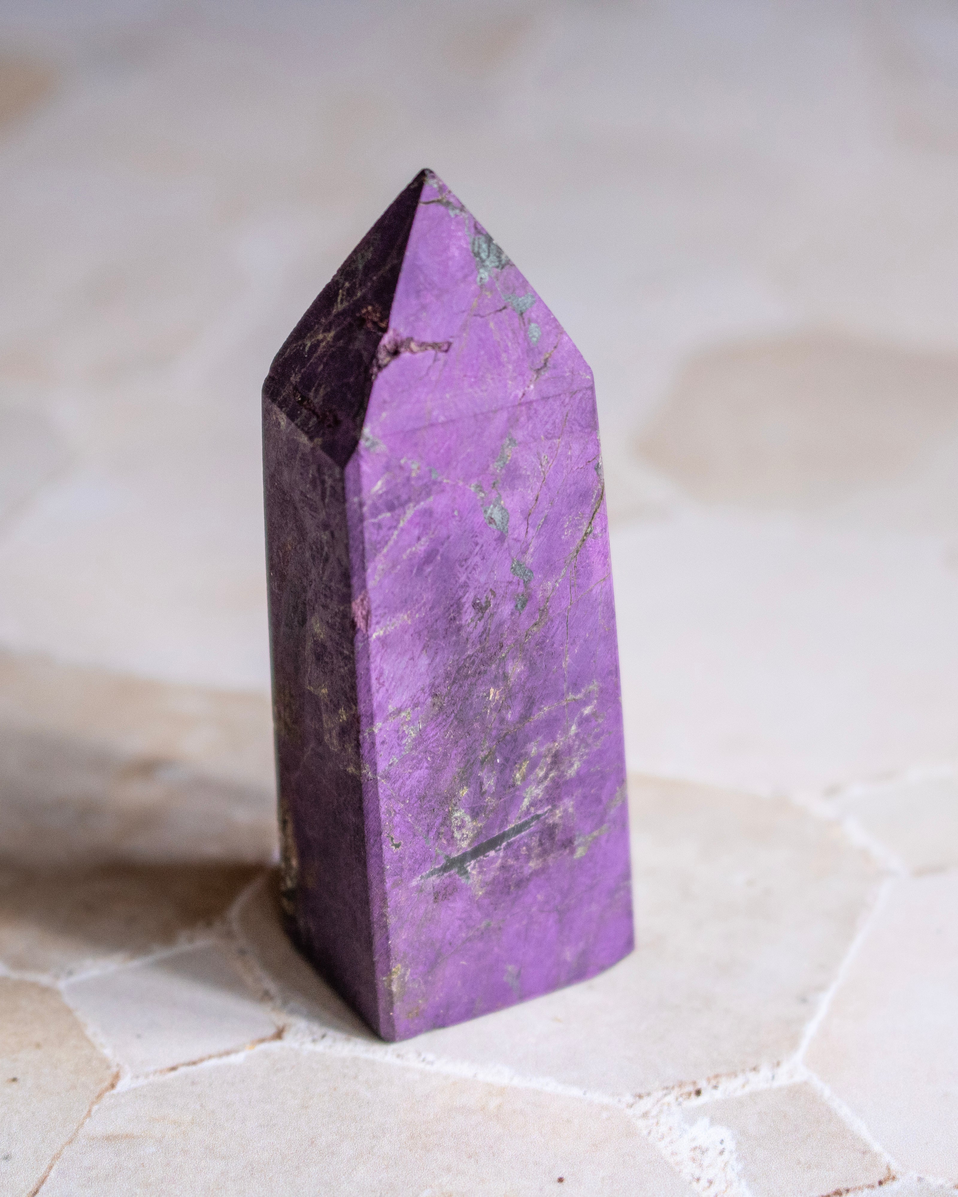 A polished purple crystal tower point with natural marbling, standing upright on a light beige stone surface.
