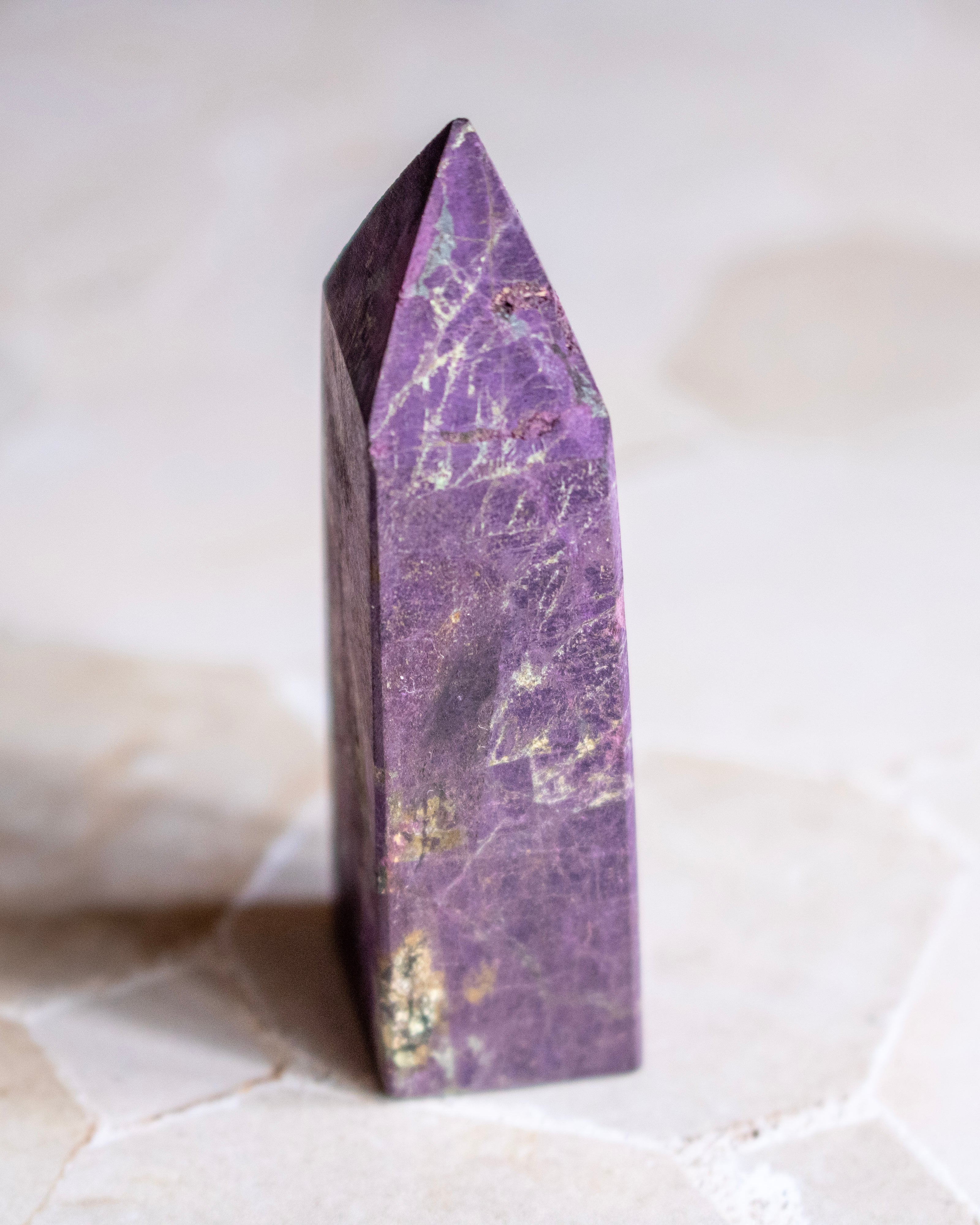 A polished purple crystal tower with natural gold and white veining, standing upright on a light beige stone surface.