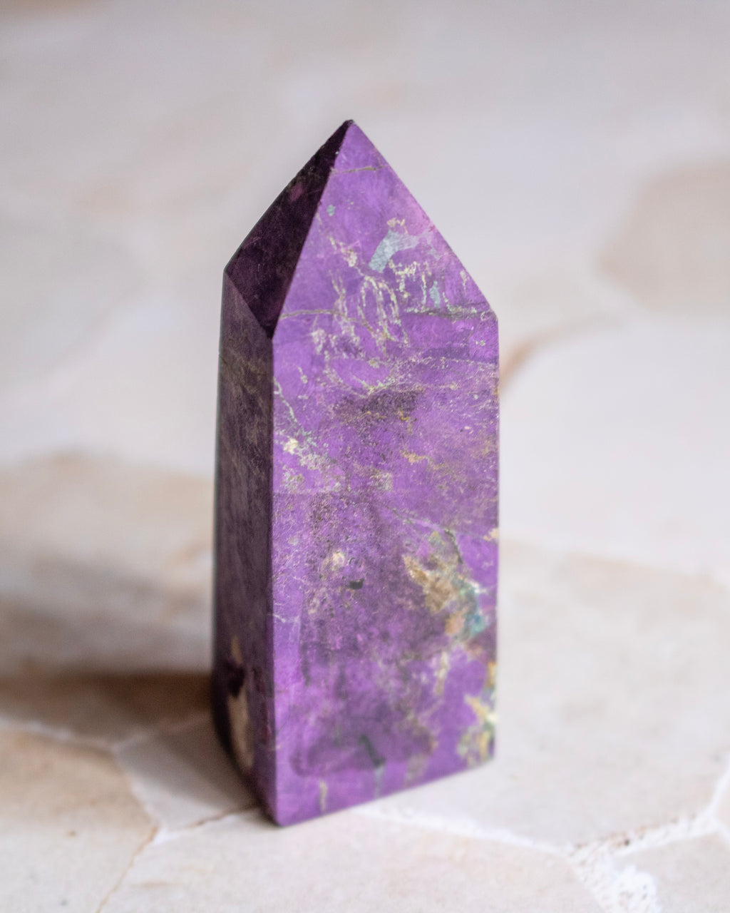 A purple amethyst crystal tower with natural gold and green matrix inclusions, standing upright on a light beige stone surface.
