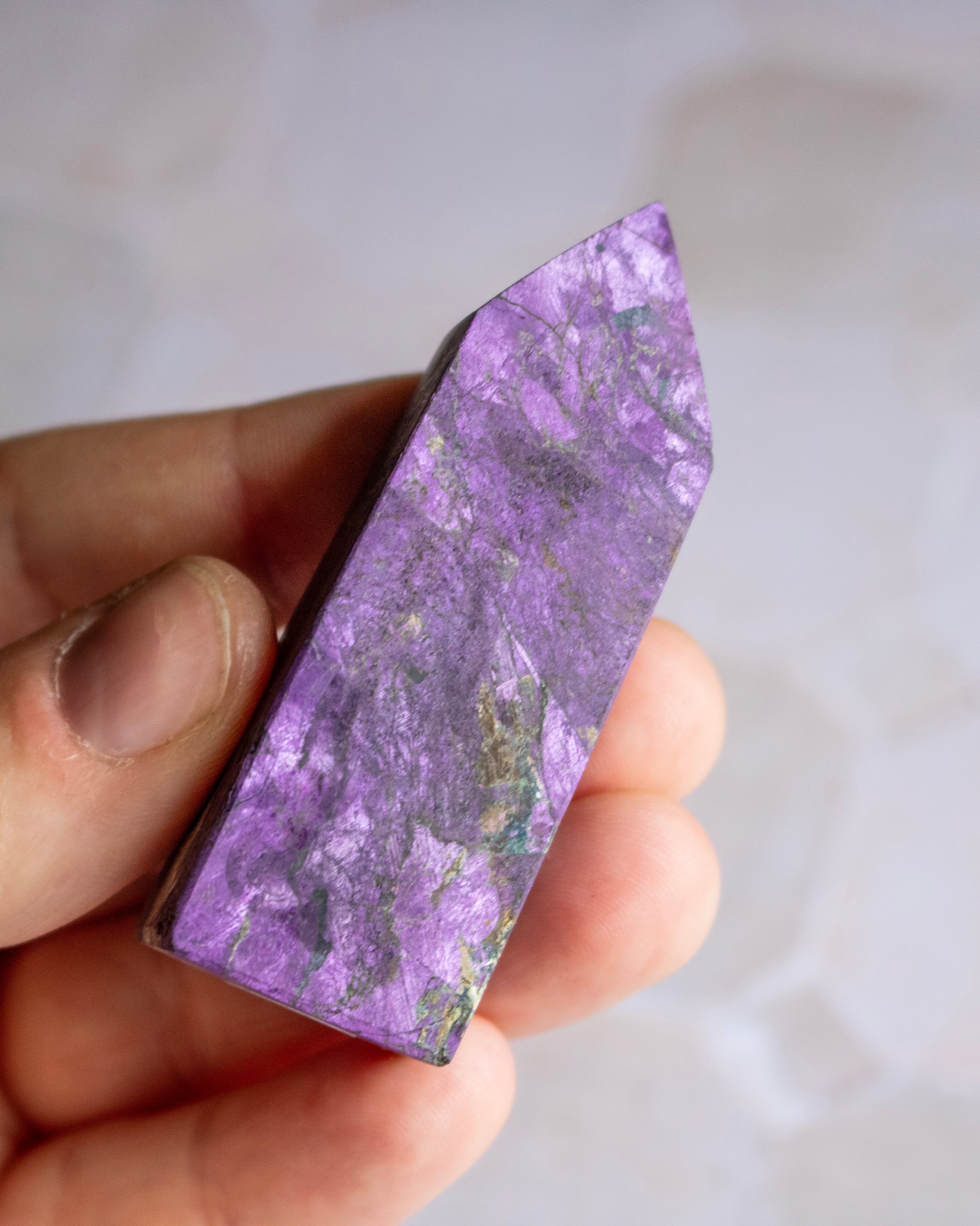 A hand holding a polished purple crystal point with natural marbled patterns of lavender, violet, and light green, set against a soft, out-of-focus light gray background.