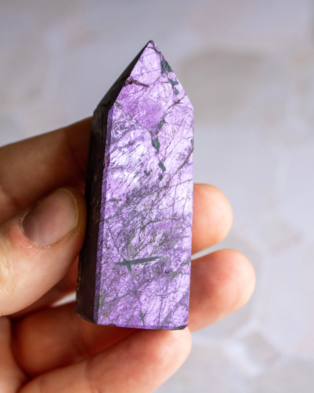 A hand holding a polished purple crystal point with natural white and dark veining, set against a soft, out-of-focus light gray background.