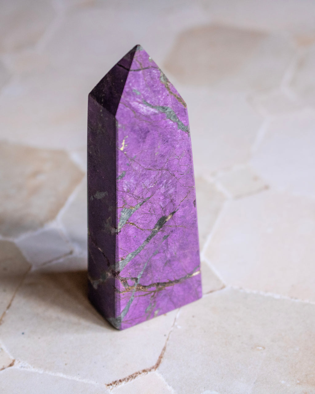 A purple jasper crystal tower with natural green and gold veining, standing upright on a light beige stone surface.