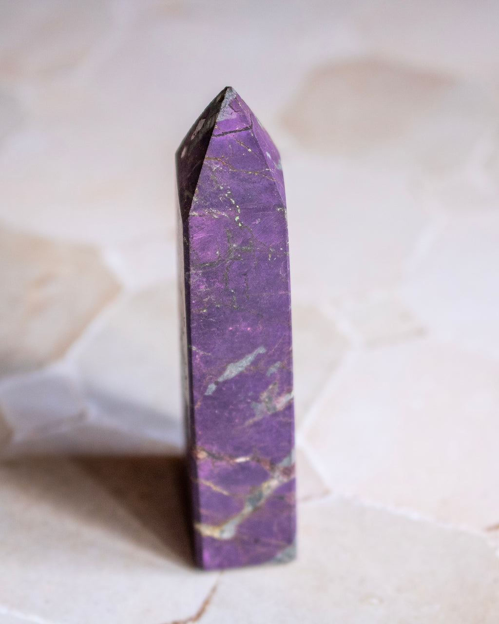A polished purple crystal point, standing upright on a light beige stone surface with a soft-focus background.