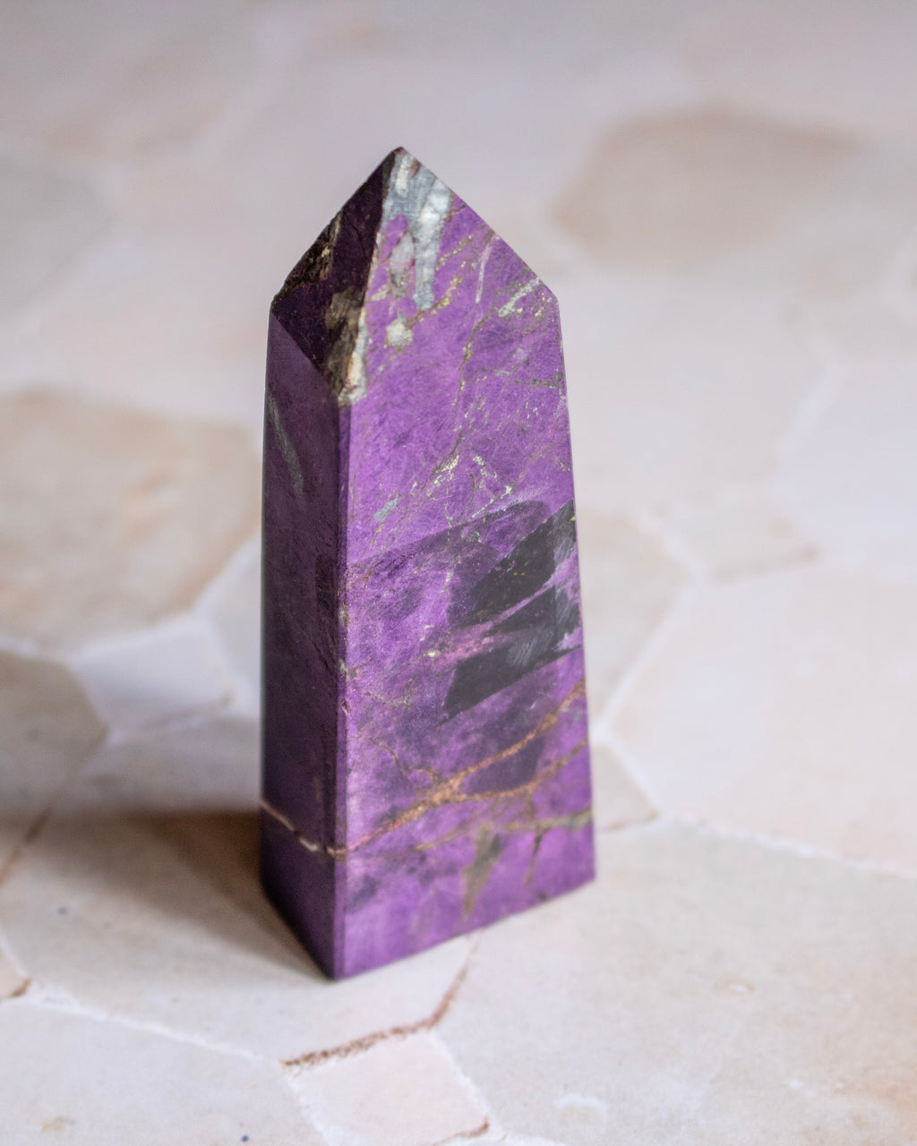 A polished purple crystal tower with natural gold and dark veining, standing upright on a light beige stone surface.