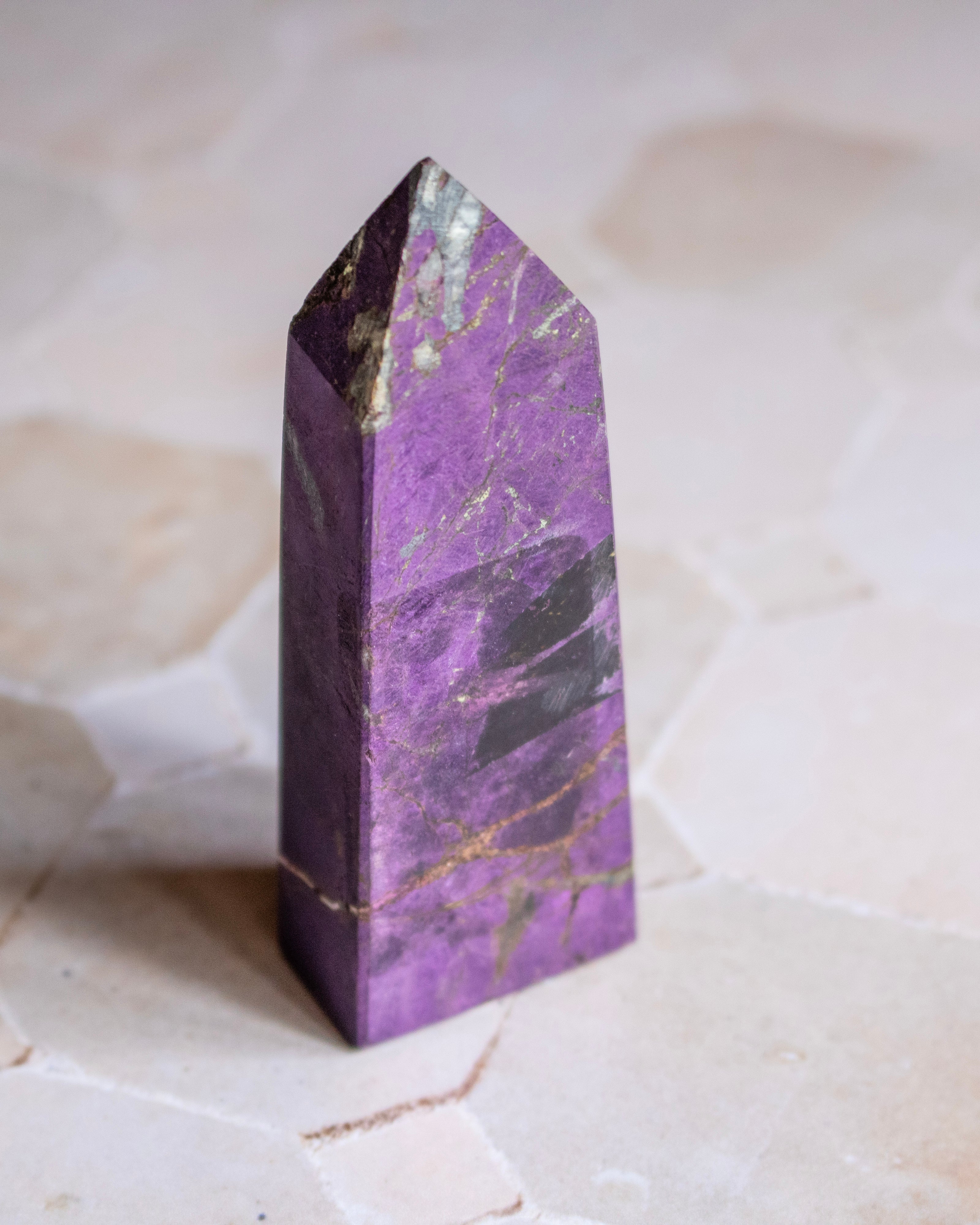A polished purple crystal tower with natural gold and dark veining, standing upright on a light beige stone surface.