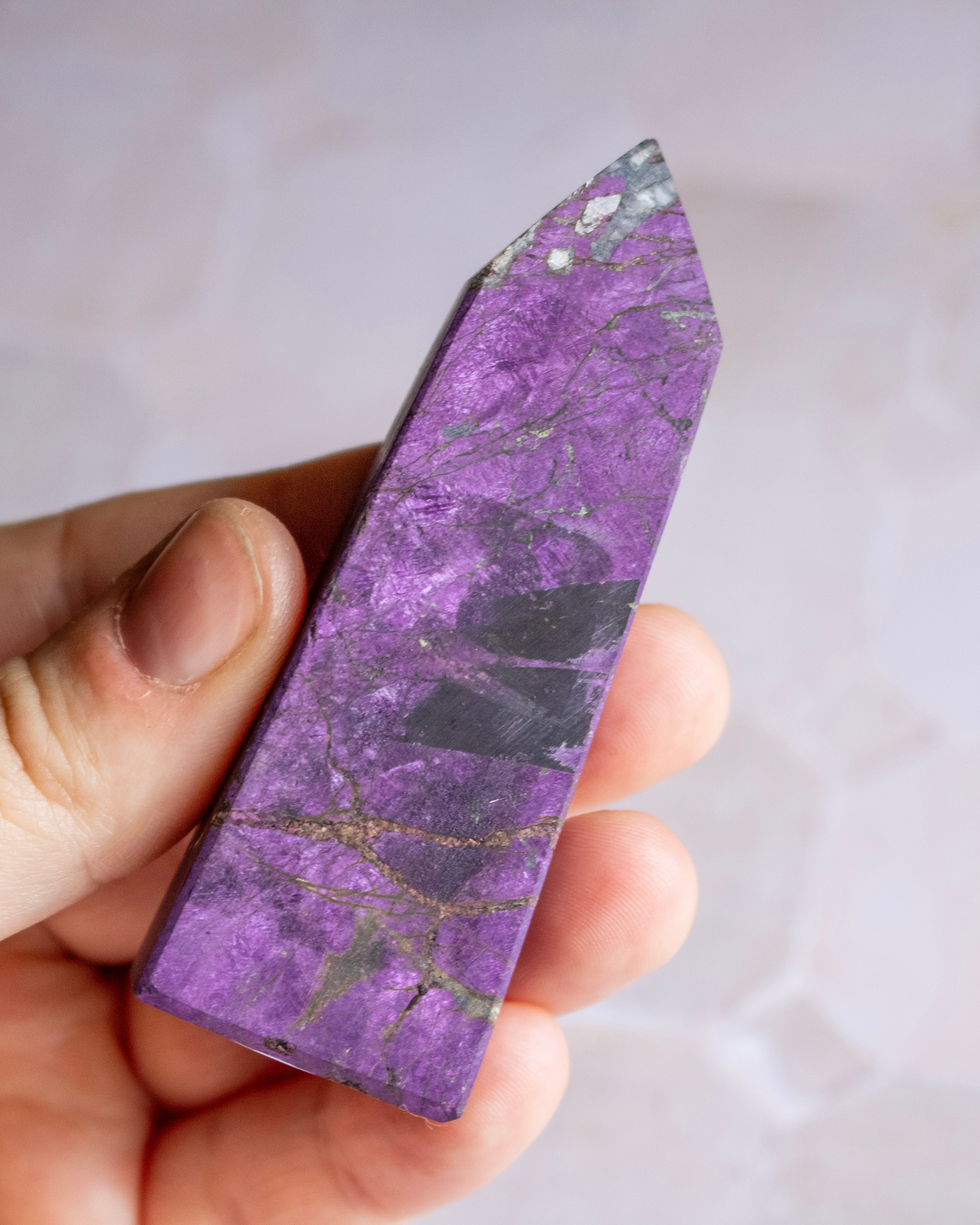 A hand holding a polished purple crystal point with natural marbling and gold veining, set against a soft, out-of-focus light background.