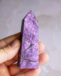 A pointed purple crystal tower with natural green and gray veining, held in a person's hand against a soft, light gray background.
